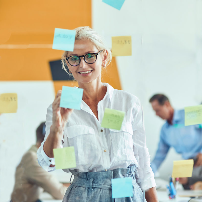 Sharing ideas. Two happy female coworkers putting colorful sticky notes on a glass wall and smiling while standing in the modern office, business people working together